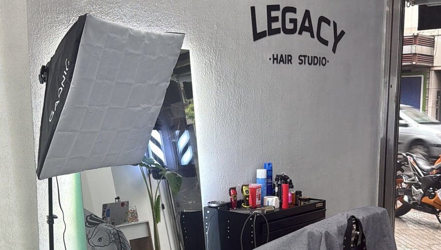 Interior de Legacy Hair Studio junto a la vidriera abierta a Avenida Ovidio Lagos, con el logo en la pared de microcemento, el softbox armado y la mesada de productos