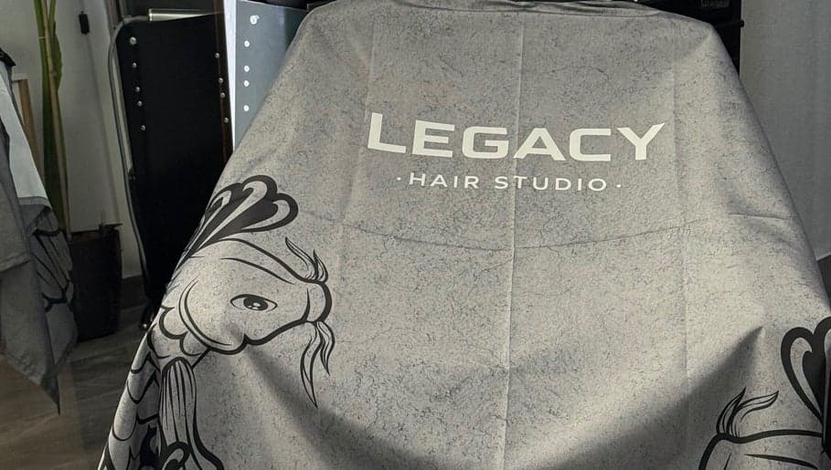 Detalle de la capa de corte de Legacy Hair Studio extendida sobre el sillon, con el logo y el koi en line-art impresos sobre la tela gris jaspeada