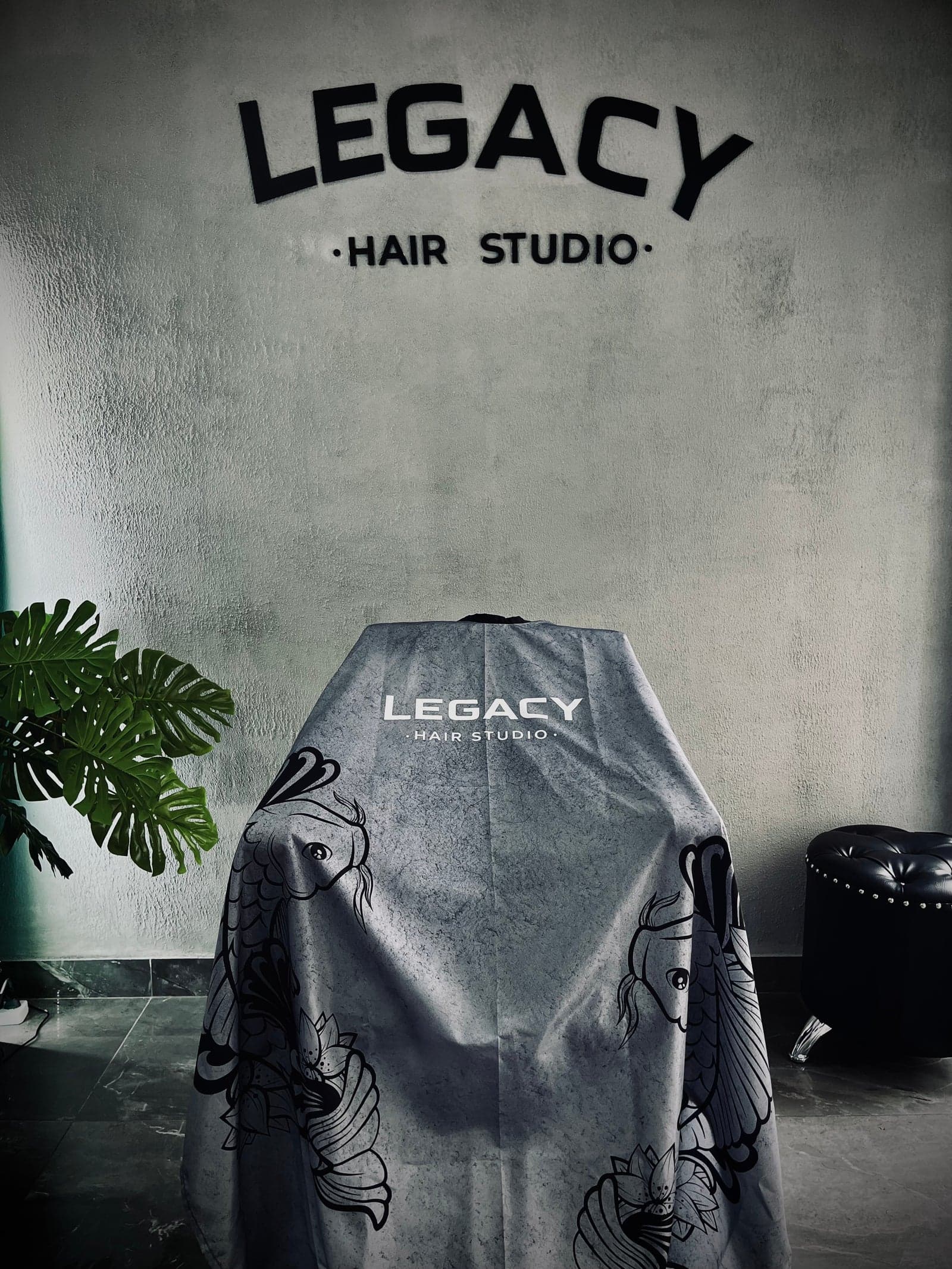 El salón de Legacy Hair Studio: el logo en relieve sobre la pared de microcemento, un barbero atendiendo frente al espejo con luz LED y el softbox encendido