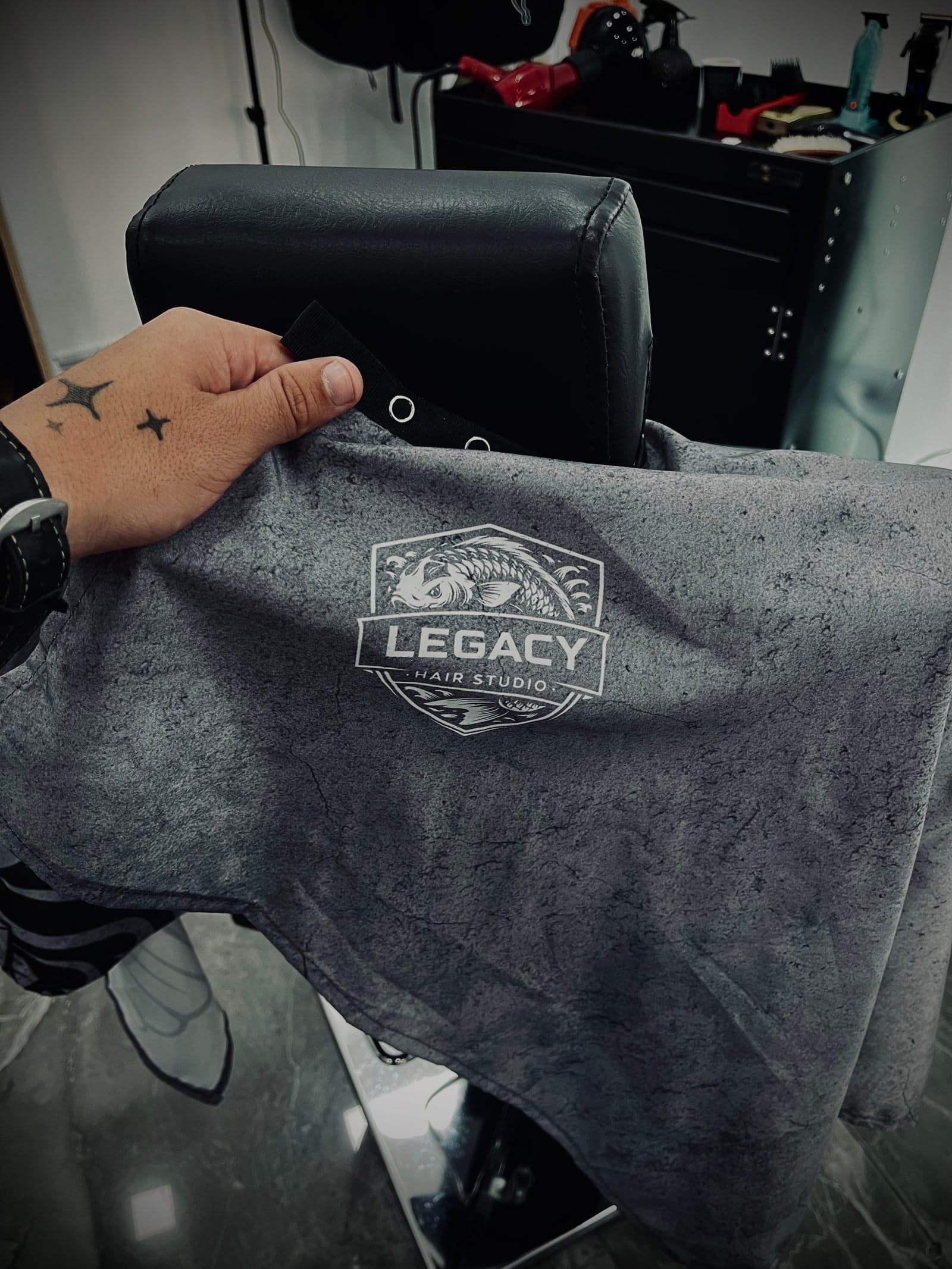 Mano de un barbero de Legacy Hair Studio sosteniendo la capa gris con el escudo hexagonal de koi bordado en el centro