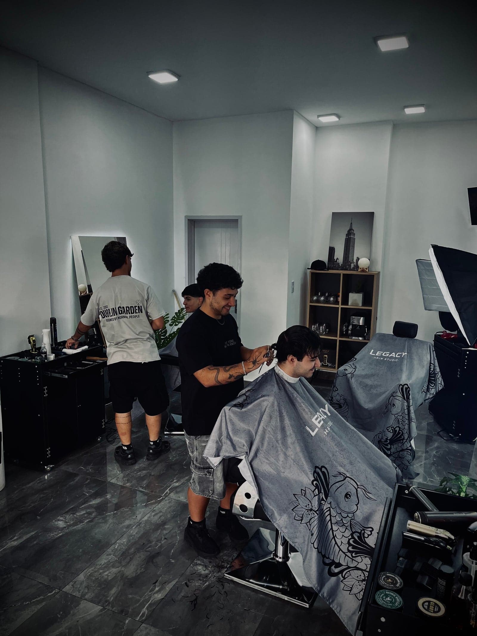 Plano general del salon de Legacy Hair Studio con un barbero cortando con tijera a un cliente sonriendo, el softbox a la derecha y el piso de marmol oscuro