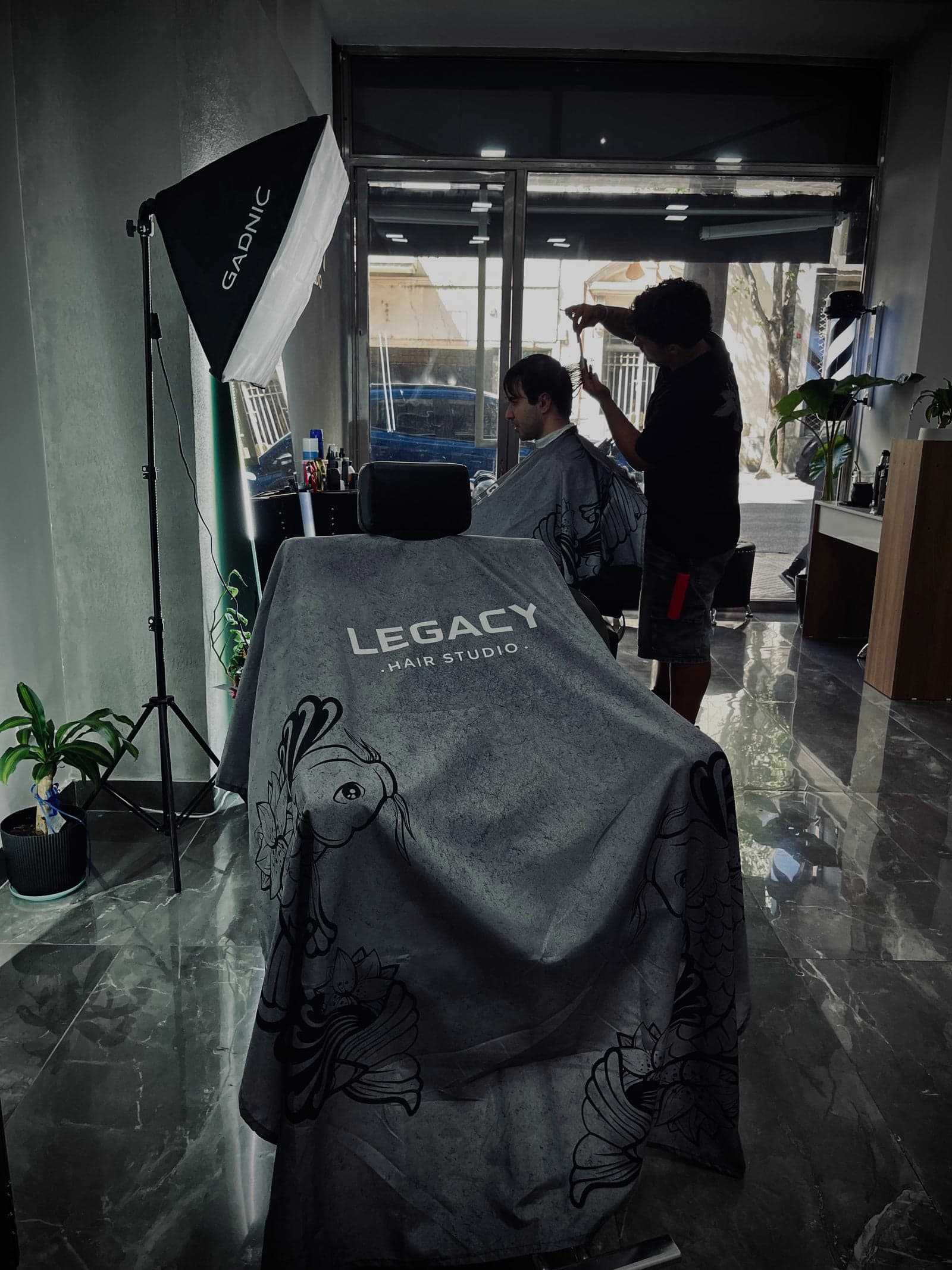 Cliente sentado en el sillon de Legacy Hair Studio con la capa de koi mientras el barbero le corta, iluminado por el softbox y con la vidriera a la calle de fondo