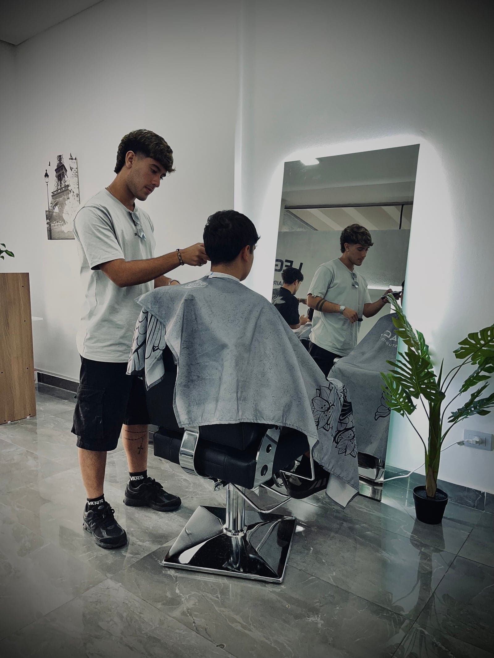 Barbero de Legacy Hair Studio perfilando la nuca de un cliente con maquina, reflejado en el espejo de cuerpo entero con marco de luz LED