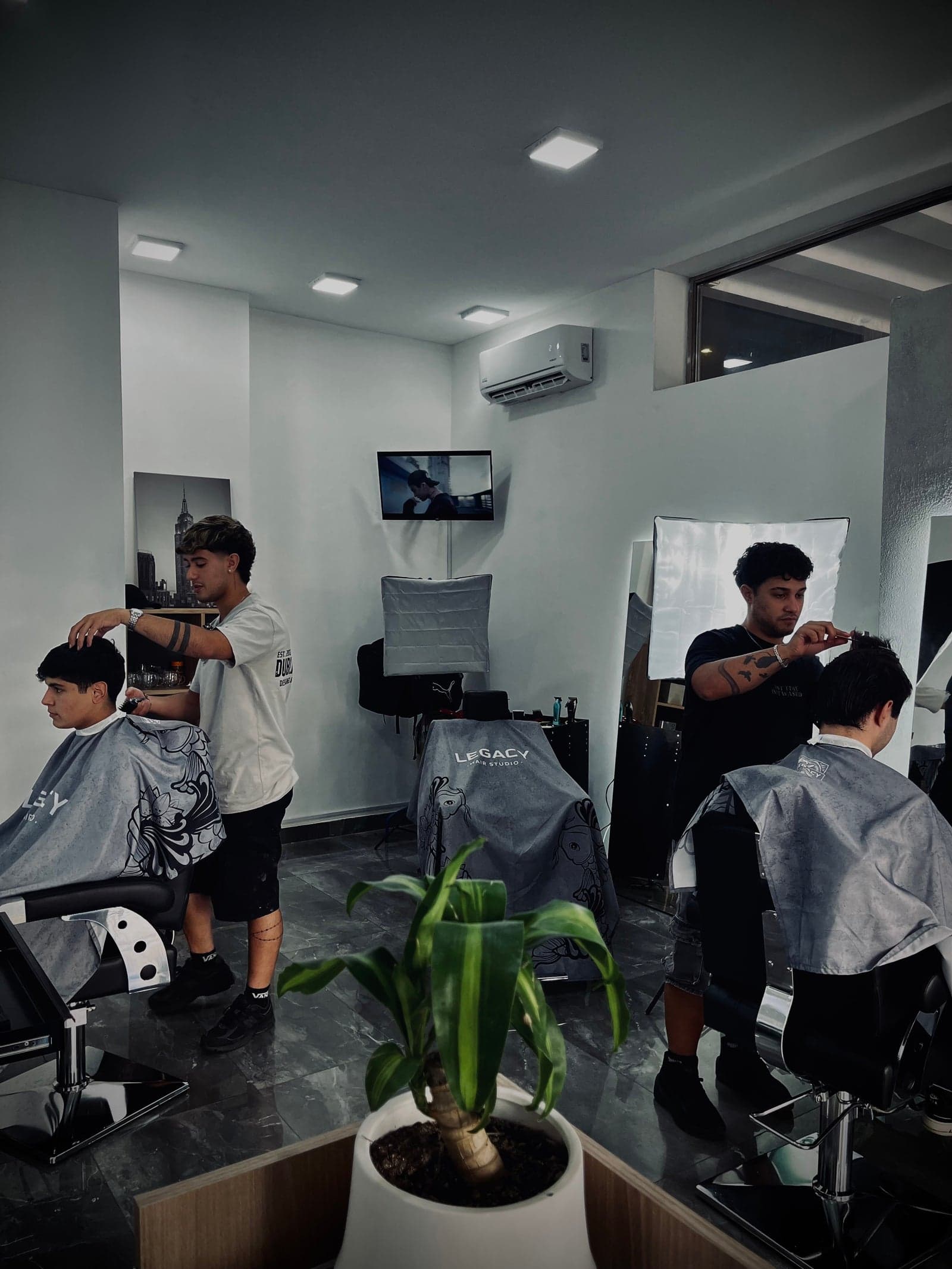 Los dos puestos de Legacy Hair Studio ocupados al mismo tiempo, dos barberos atendiendo clientes con las capas de koi bajo la luz de los softbox